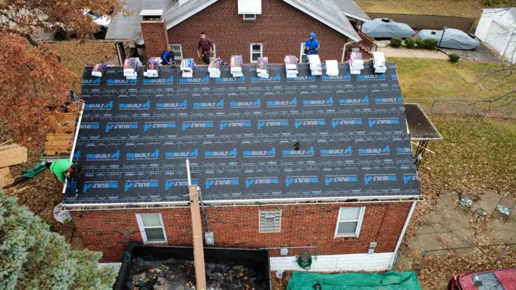 An overhead view of a roof being worked on, with several workers installing roofing materials. The roof is covered with black underlayment and edged with stacks of shingles. The house is a brick structure with a tan and white color scheme.