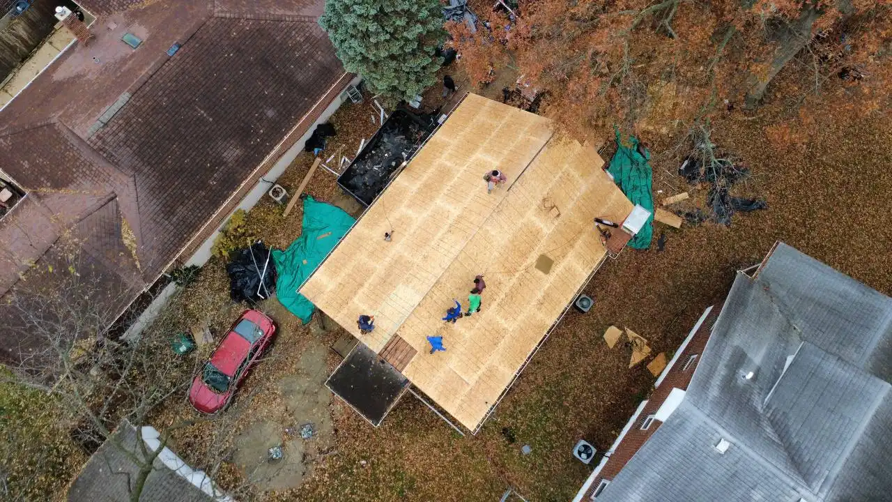 An aerial view of a construction site where workers are installing a new roof on a house. Surrounding trees have autumn foliage, and various construction materials are laid out on the ground, including tarps and wood planks.