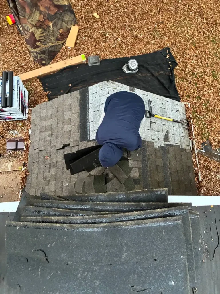 A person in a blue hoodie is working on a roof, laying down shingles, surrounded by a blanket of autumn leaves.