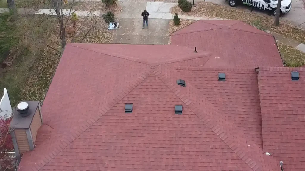 An aerial view of a residential roof with a reddish-brown surface and several vents. In the background, a person is standing on the driveway, surrounded by autumn leaves, while a vehicle is parked nearby.