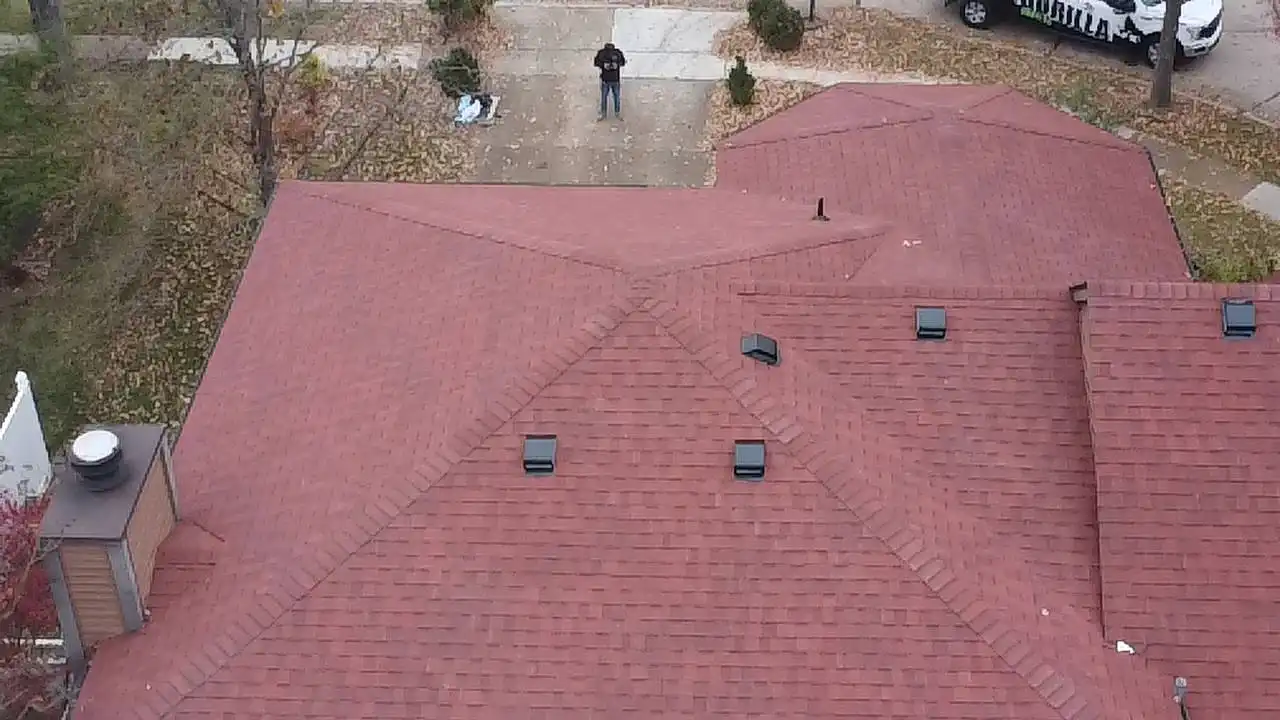 An aerial view of a residential roof with a reddish-brown surface and several vents. In the background, a person is standing on the driveway, surrounded by autumn leaves, while a vehicle is parked nearby.