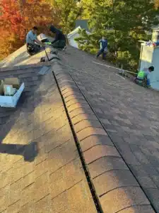Two workers installing roofing materials on a sloped roof, with trees displaying autumn foliage in the background.