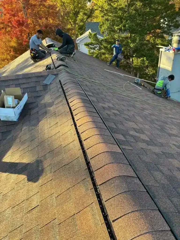 Two workers installing roofing materials on a sloped roof, with trees displaying autumn foliage in the background.