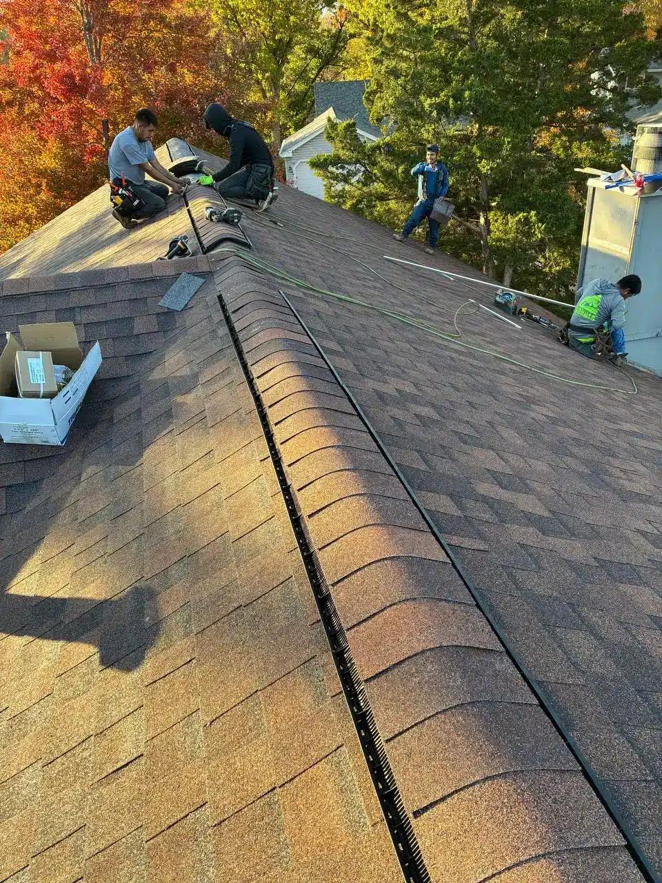 Two workers installing roofing materials on a sloped roof, with trees displaying autumn foliage in the background.