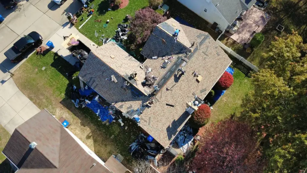 An aerial view of a residential house undergoing roof repairs, with multiple workers on the roof and scattered materials around the yard.