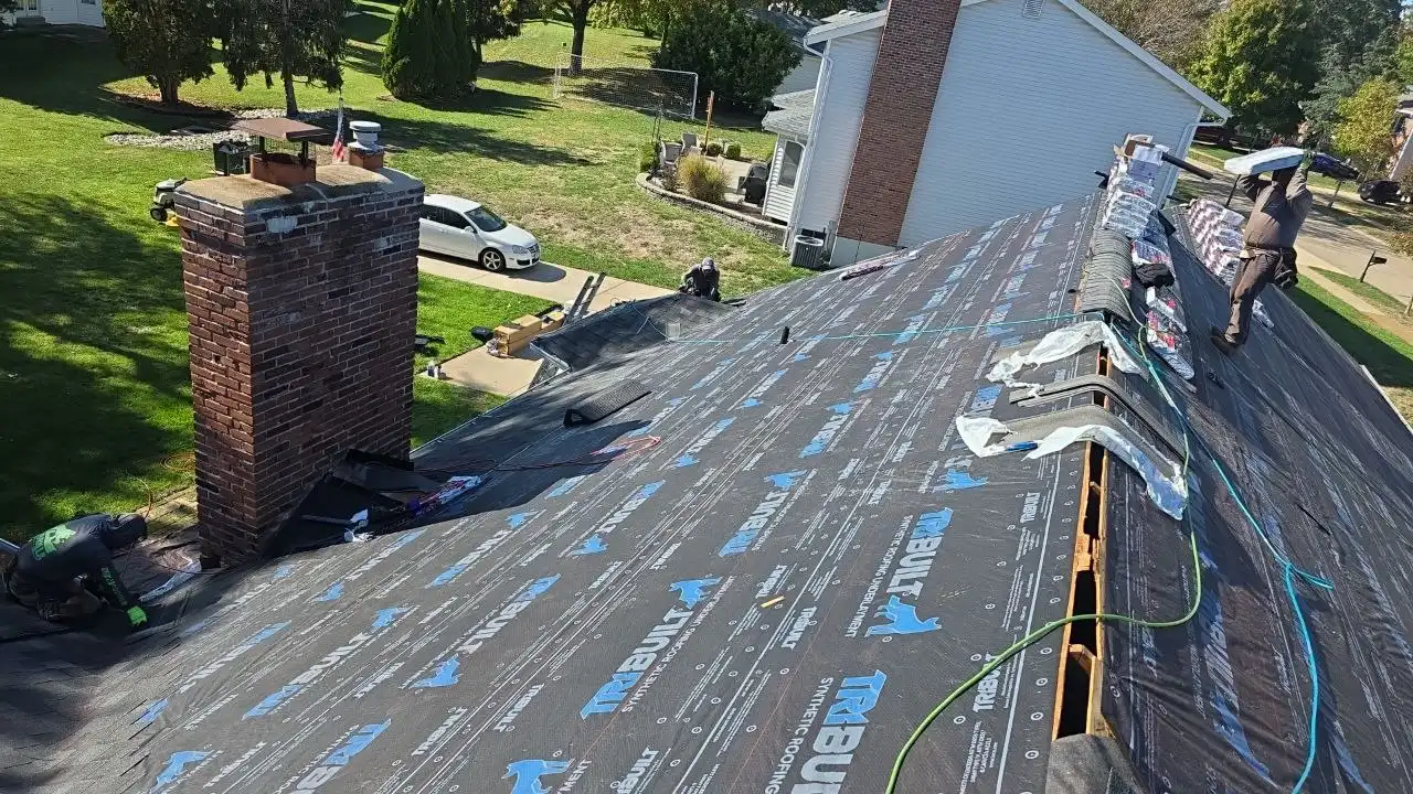 roofing-installation-process Workers laying roofing material on a sloped roof, with a chimney in view and a clear green lawn below.