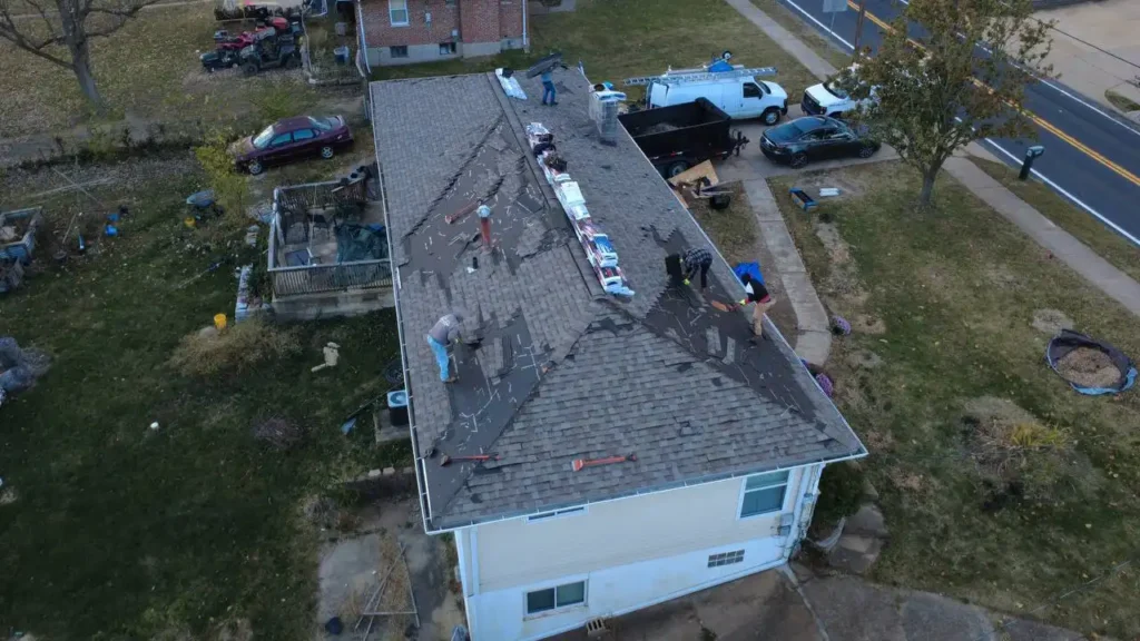 Aerial view of workers repairing a roof on a single-story house. They are removing old shingles and preparing for new roofing materials, with tools scattered around. Nearby, there are several vehicles parked on the lawn and along the road.