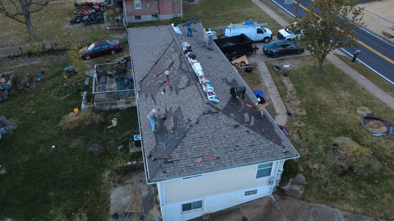 Aerial view of workers repairing a roof on a single-story house. They are removing old shingles and preparing for new roofing materials, with tools scattered around. Nearby, there are several vehicles parked on the lawn and along the road.