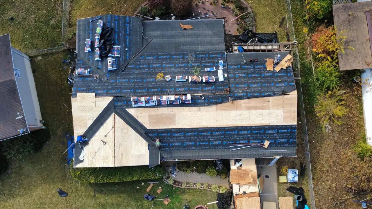 Aerial view of a house under renovation, showing a partially completed roof with exposed plywood and roofing materials scattered on top, including insulation and rolls of roofing felt.