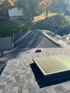 A rooftop view showing a person working on a gray shingle roof. The scene is illuminated by sunlight, with colorful autumn trees in the background and a grassy lawn below.