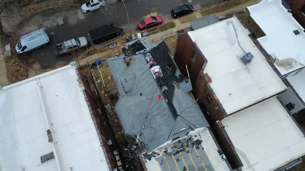 An aerial view of a residential area showing several houses with different roofing materials. One house has workers on the roof, surrounded by various construction tools and materials.