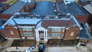 An aerial view of a brick building undergoing roof repairs. The roof on the left is partially covered with blue tarpaulin, and workers can be seen making updates. In the foreground, construction materials and debris are visible, along with a ladder and a trash container.