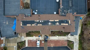 An aerial view of a building undergoing roof repairs, featuring workers on the roof and equipment scattered around. The structure shows patched areas and a circular driveway leading to a parked truck.