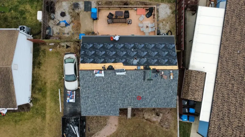 An aerial view of a house under roof renovation, showing workers on the roof, a wooden deck with outdoor furniture, and a parked car nearby.