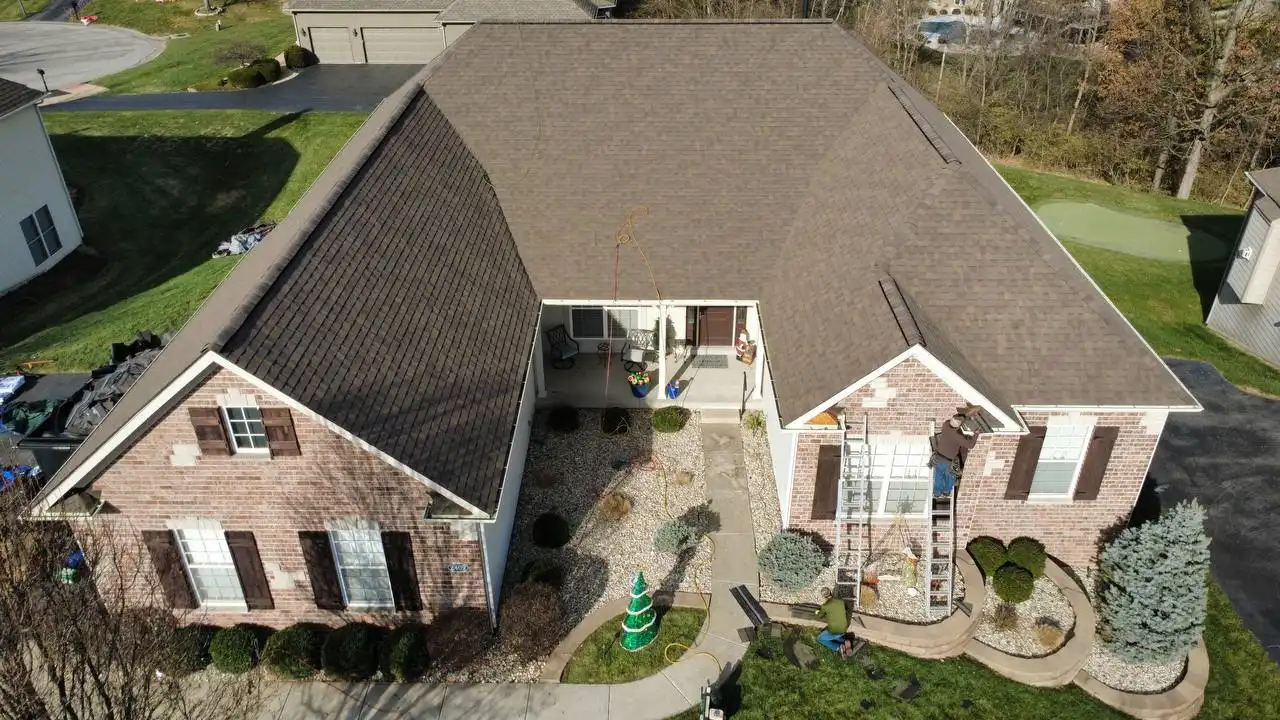 Aerial view of a brick house with a brown roof, surrounded by a well-maintained lawn and landscaping. Two people are working on a ladder in front of the house, and there is a small decorative Christmas tree in the garden.