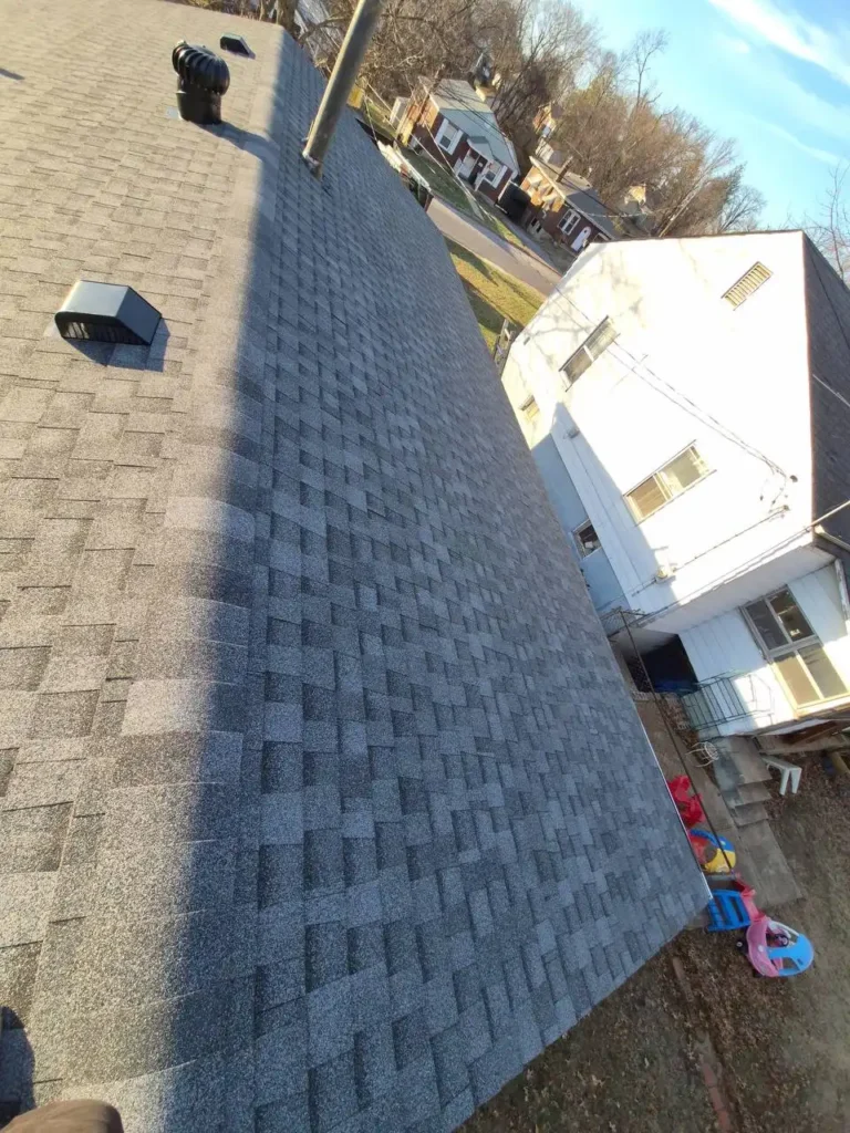 A view of a sloped roof covered with gray asphalt shingles, featuring vents and a chimney. In the background, there are several houses and trees under a clear sky.