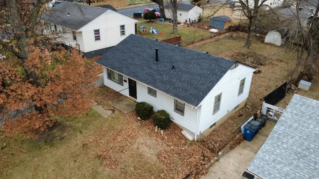 An aerial view of a small white house with a black roof, surrounded by dry leaves in a residential neighborhood. Nearby homes and a few outdoor toys are visible.