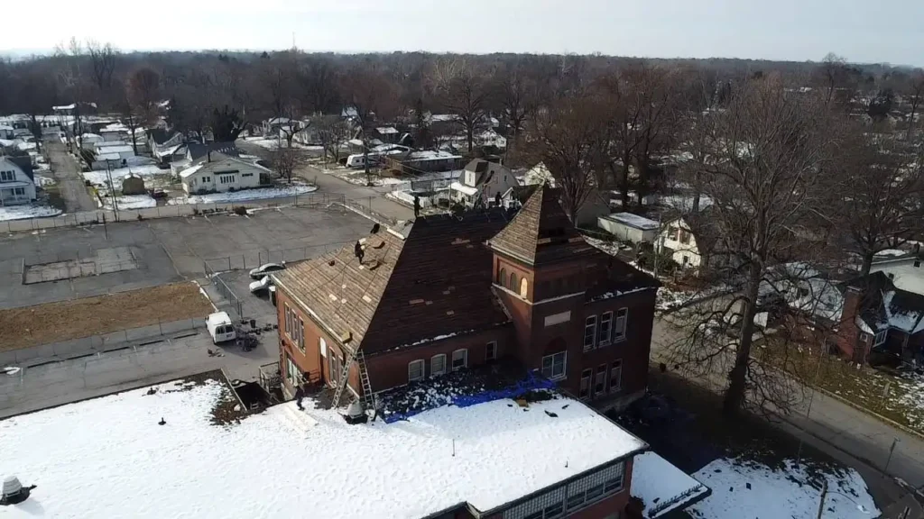 An aerial view of a large brick building with a snowy rooftop, surrounded by a neighborhood of houses and trees. Workers are seen on the roof, and a few vehicles are parked nearby.