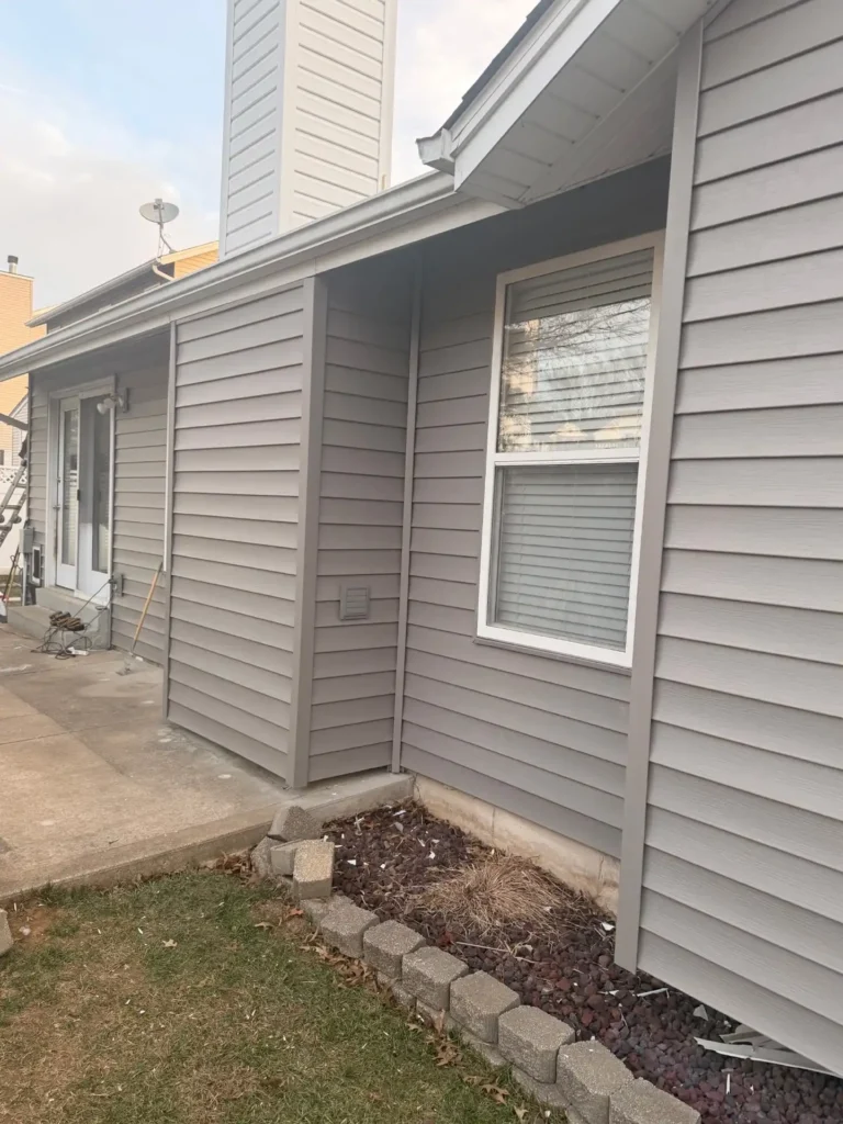 A side view of a gray house featuring horizontal siding, a chimney, and a window. The foundation has stone landscaping, and the area is tidy but shows some dry grass.