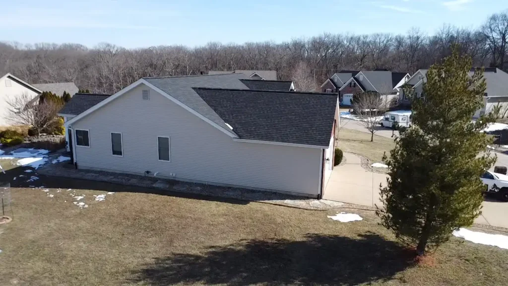 A side view of a light-colored house with a black roof, nestled in a suburban area with trees and other homes in the background. The lawn is mostly brown with patches of snow, indicating early spring.