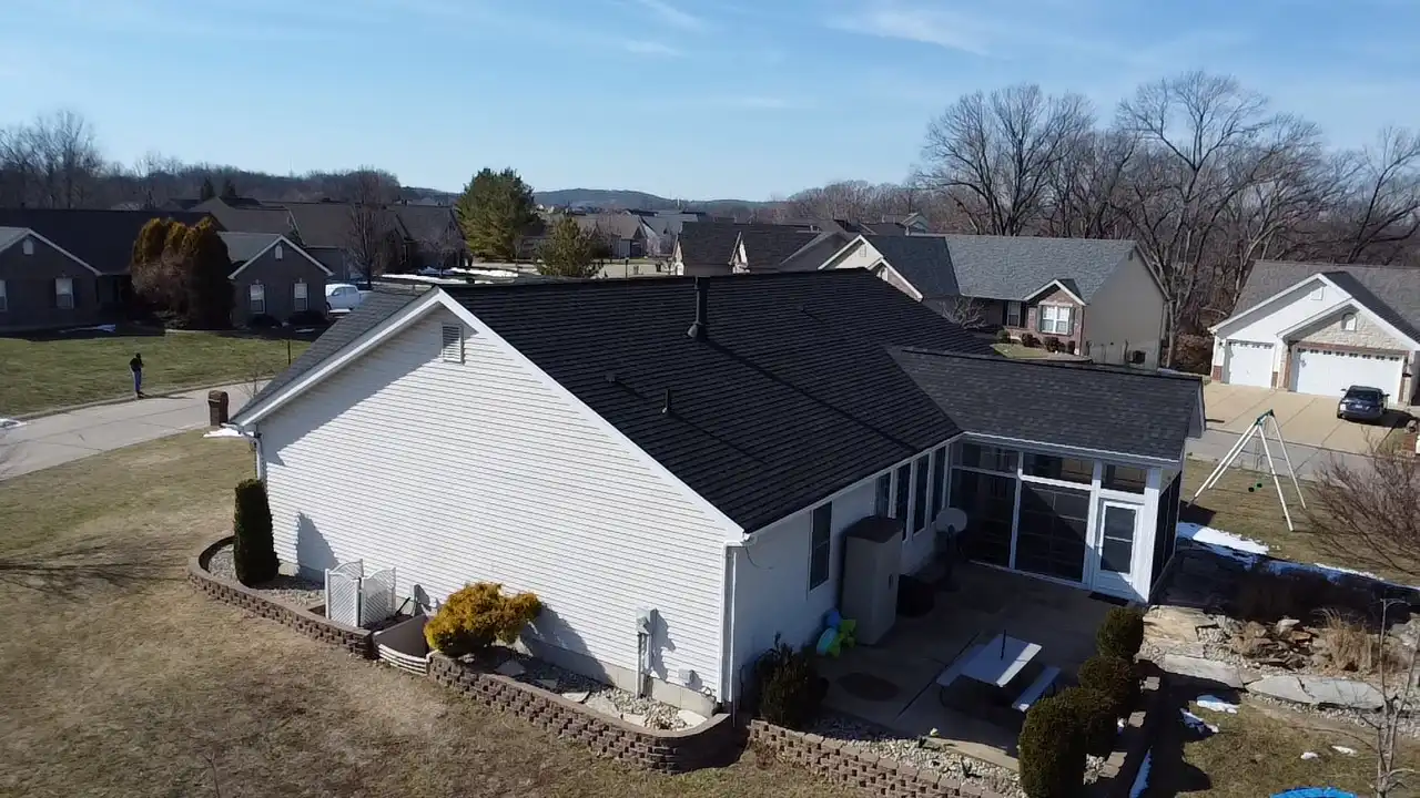 Aerial view of a residential home with a dark roof, surrounded by other houses in a suburban area, under a clear blue sky.