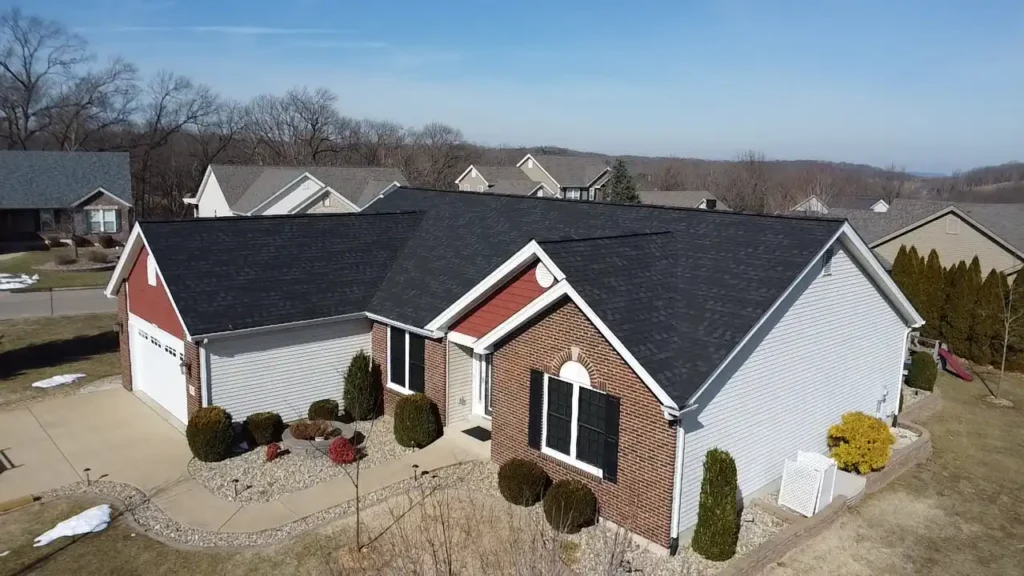 Aerial view of a beautiful suburban home with a mix of brick and siding. The house features a well-maintained lawn with decorative bushes and a pathway leading to the front door. Nearby homes are visible in the background under a clear blue sky.