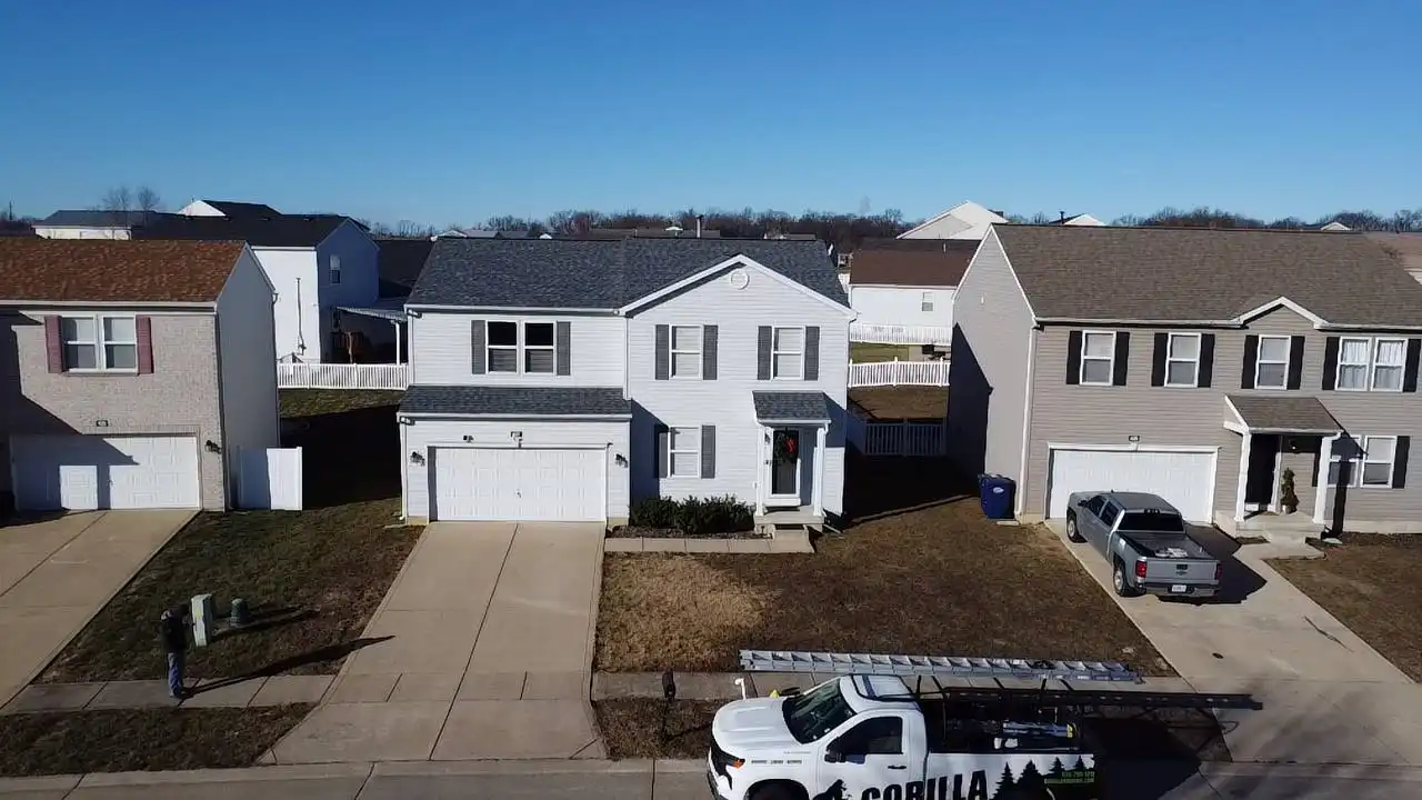 Aerial view of a two-story, light blue house with a gray roof, flanked by two other homes, on a clear day. A white truck with a ladder is parked in the driveway, while a person stands near a utility box.