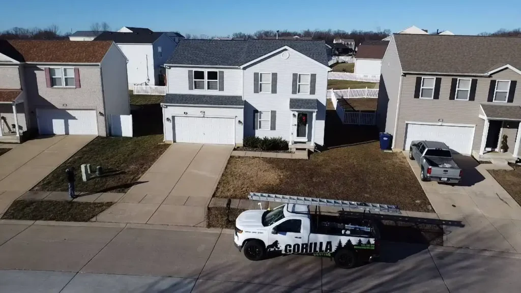 An aerial view of a suburban neighborhood featuring two houses. One house is white with gray shutters, and another is light brown. A white truck with a ladder and the word 'GORILLA' on its side is parked in front of the white house.