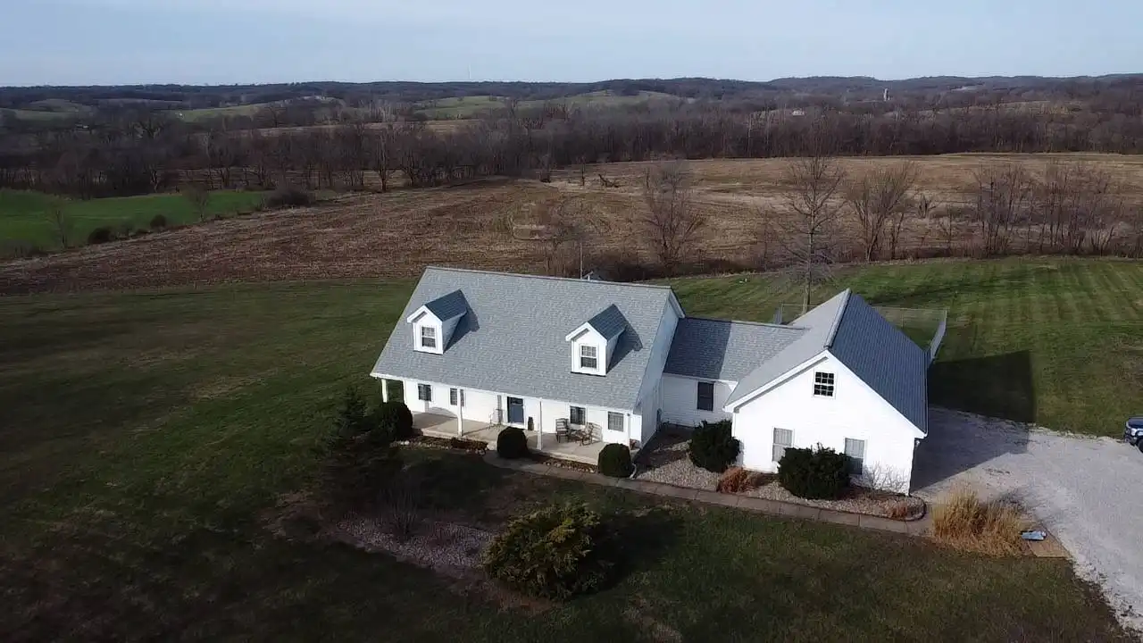 Aerial view of a white house with a grey roof, surrounded by grassy fields and trees, under a clear sky.