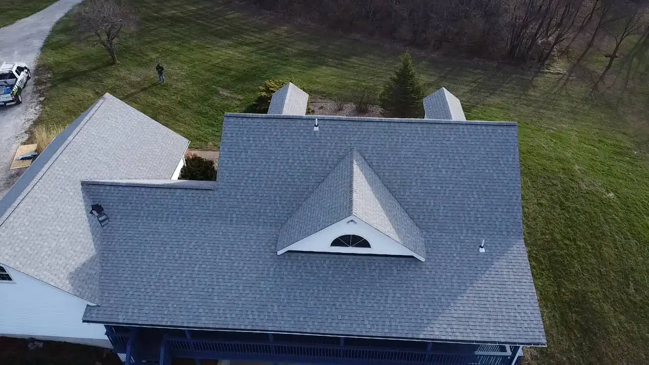 aerial-view-of-house-with-peaked-roof Aerial view of a house with a peaked roof and a white exterior, surrounded by green grass and a few trees. A person can be seen in the distance near a parked vehicle.